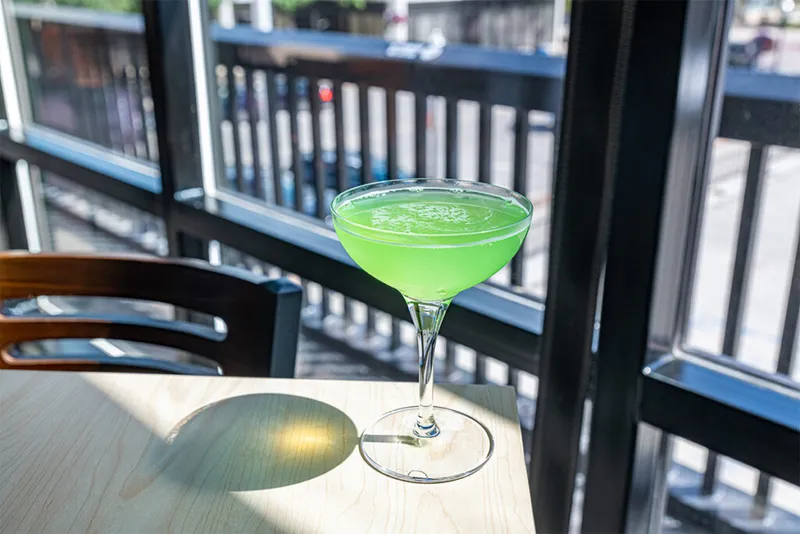 Bright green cocktail in a coupe glass on a light wood table by sunlit windows