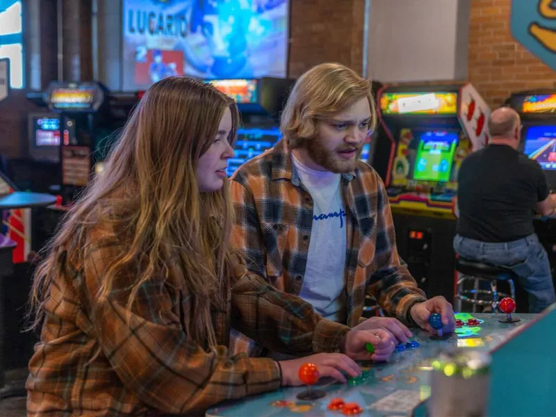 Two young adults in flannel shirts playing an arcade game in a retro bar arcade