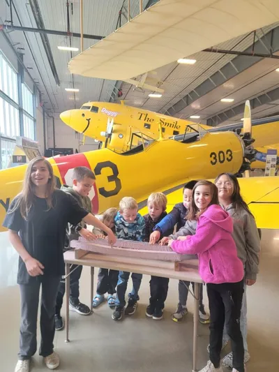 Children gathered around a table in an aviation museum hangar with yellow vintage aircraft behind them