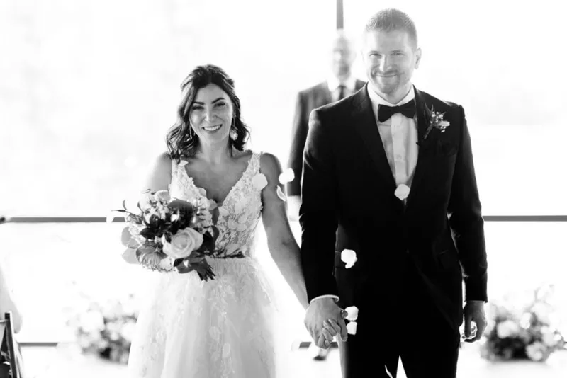 Smiling bride in lace gown holding bouquet walks hand-in-hand with groom in black tuxedo