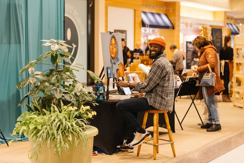 Smiling artist in orange beanie painting a portrait on an easel at an indoor craft fair