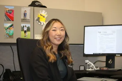 Smiling woman in a blazer seated at an office desk with a monitor and pinned photos behind her