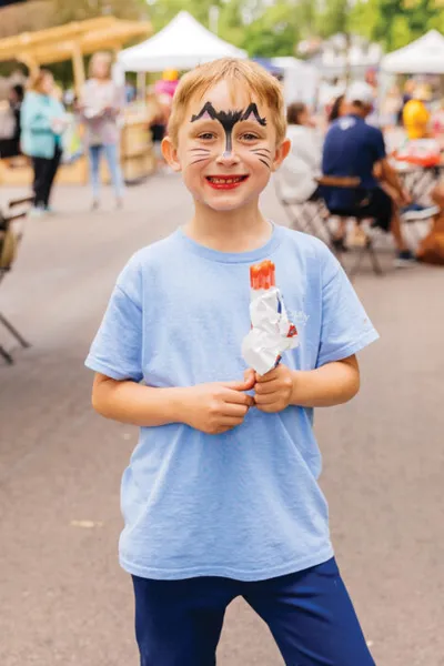 Smiling boy with cat face paint holds an icy treat at an outdoor community event