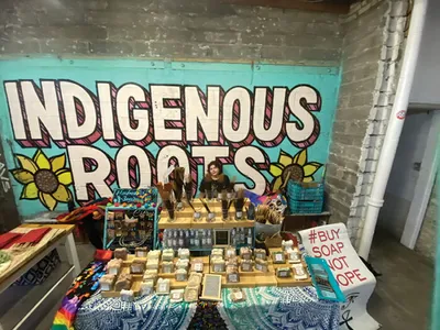 Vendor stands behind a table of handmade soaps beneath a teal "Indigenous Roots" mural with sunflowers