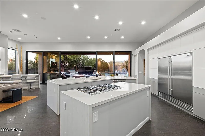 Modern open-plan kitchen with two white islands, gas cooktop, stainless steel fridge, and floor-to-ceiling windows