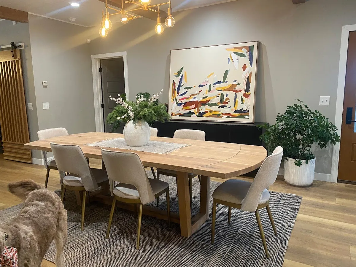 Modern dining room with a live-edge wood table, upholstered chairs, abstract wall art, and a curly dog in the foreground