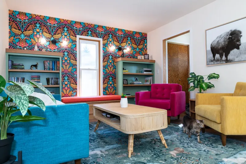 Colorful living room with bold floral wallpaper, teal bookshelves, magenta and yellow chairs, rattan coffee table, and a cat