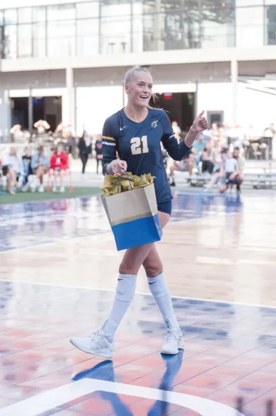 Smiling volleyball player in navy number 21 jersey carries a gift bag across an outdoor court
