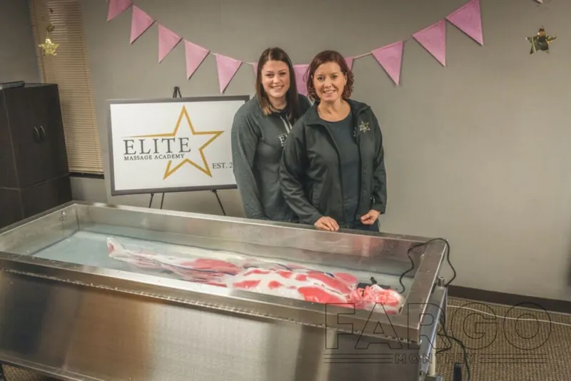 Two women in matching jackets stand behind a hydrotherapy tub near an Elite Massage Academy sign