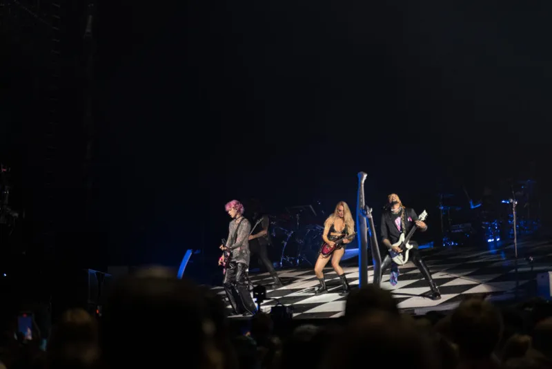 Three guitarists performing on a checkerboard stage before a dark arena crowd