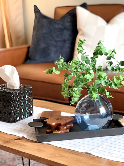 Eucalyptus stems in a round blue vase on a black tray atop a wooden coffee table, with a tan leather sofa behind