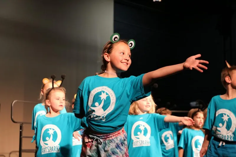 Girl in frog headband and teal ArtSpark T-shirt performs on stage with other children