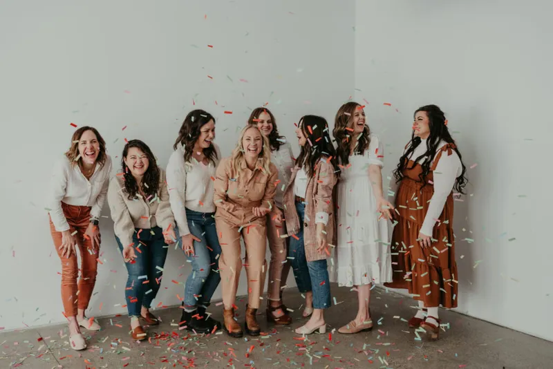 Seven laughing women pose together as colorful confetti rains down in a bright studio