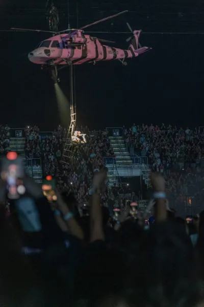 Pink striped helicopter hovers over a packed arena crowd as a performer dangles from a ladder below