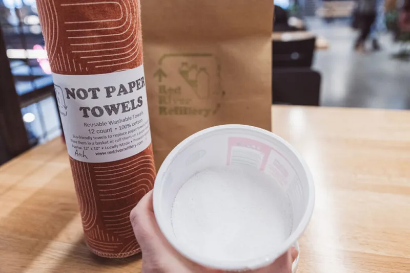 Hand holding a foam cup beside a roll of reusable cotton towels labeled 'Not Paper Towels' on a wooden table