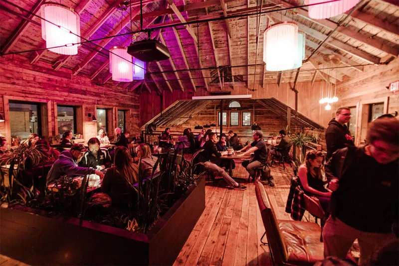 Crowded rustic bar interior with wood-plank ceiling, vaulted rafters, and pink ambient lighting