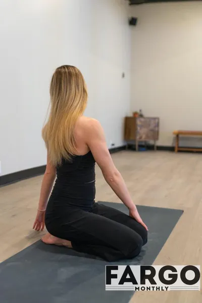 Woman in black activewear seated on a gray yoga mat facing away in a bright studio