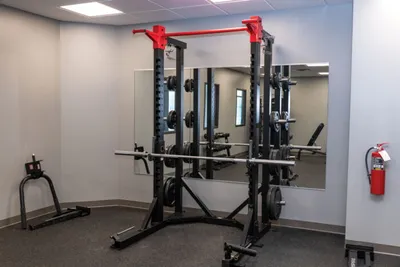 Black and red power rack with barbell in a mirrored gym room with a fire extinguisher on the wall