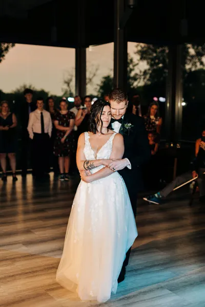 Bride and groom share first dance, guests watching in background at dusk