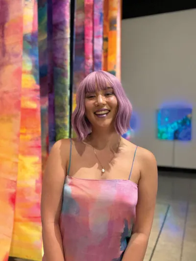 Smiling woman with lavender hair and pastel tie-dye top stands before colorful hanging textile art in a gallery