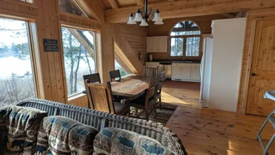 Rustic wood-paneled cabin interior with dining table, chandelier, and snowy lake view through large windows