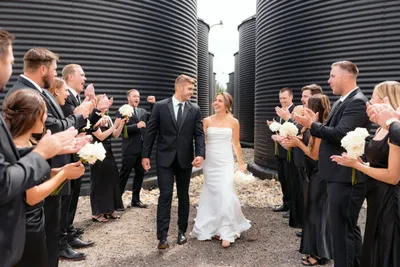 Bride and groom walk between cheering wedding party flanked by large black industrial silos