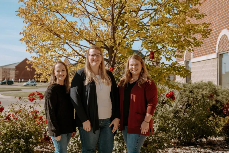 Three smiling women standing outdoors in front of a tree with golden fall foliage