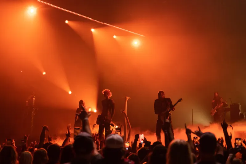 Rock band performs on a fog-filled stage bathed in orange light before a packed crowd