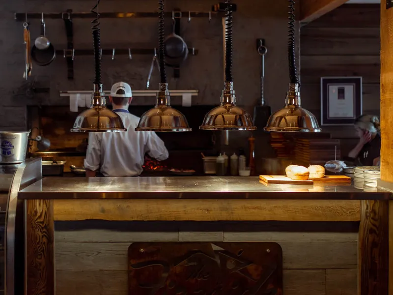 Cook in white jacket and cap works at an open kitchen pass under polished pendant lamps