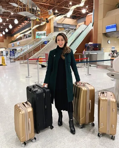 Smiling woman in a dark green coat standing with three suitcases in an airport terminal