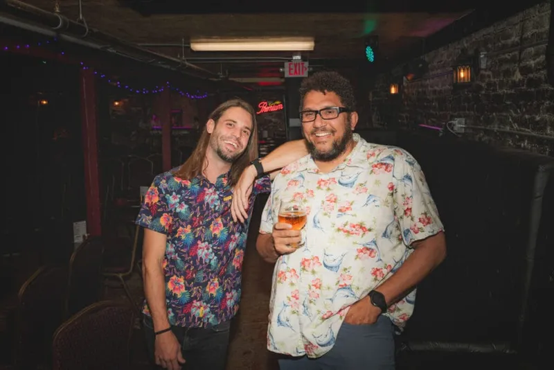 Two smiling men in floral shirts pose together inside a dimly lit bar, one holding a beer
