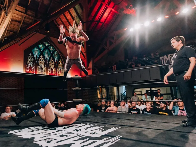 Shirtless wrestler leaps off ropes toward a fallen opponent in a converted church venue with stained-glass windows
