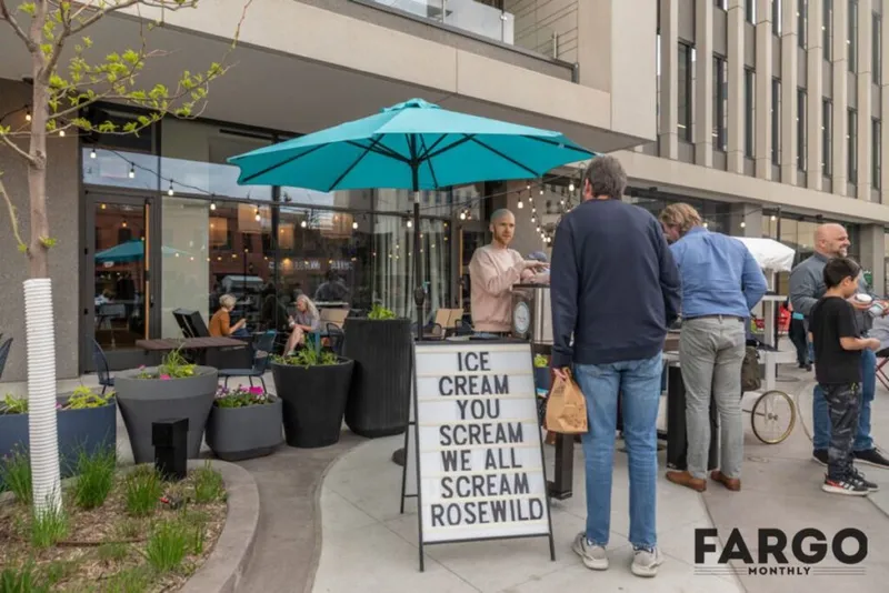Customers line up at an outdoor ice cream stand under a teal umbrella beside a sign reading 'Rosewild