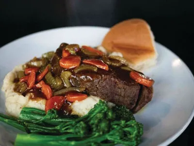 Braised beef over mashed potatoes with sautéed peppers, carrots, and broccoli rabe alongside a dinner roll