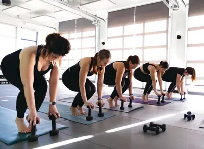 Women in black activewear doing a barre workout with small dumbbells on mats in a bright studio