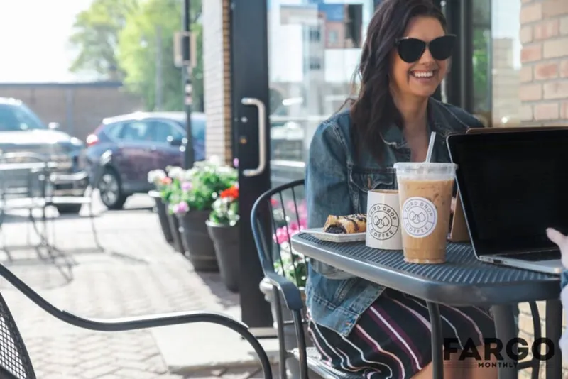 Smiling woman in sunglasses and denim jacket at a sunny café patio with iced coffee and a laptop