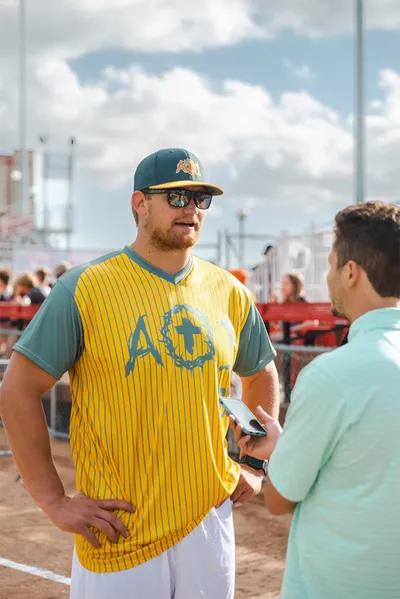 Baseball player in yellow pinstripe AO1 jersey and green cap speaks with interviewer outdoors