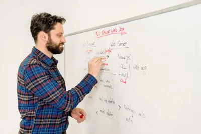 Bearded man in plaid shirt writing on a whiteboard with a red marker