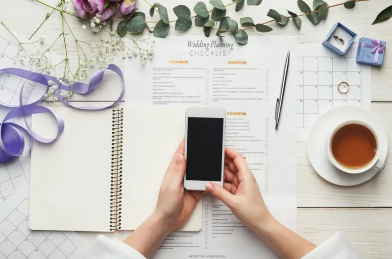 person holding their phone with a wedding checklist and calendars on the desk in front of them