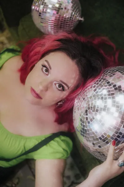 Woman with red hair in a green top lying down holding a disco ball