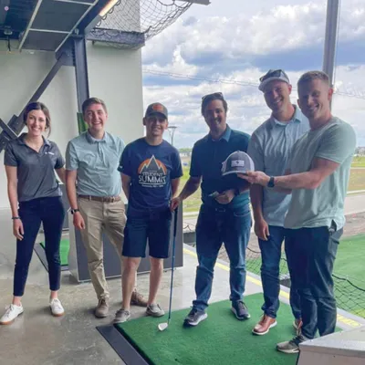 Six adults smiling at a golf driving range bay, two holding a baseball cap