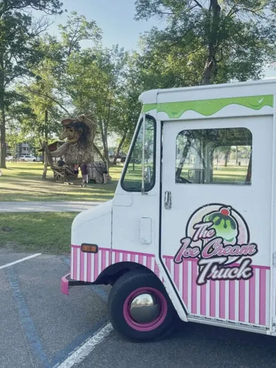 White and pink ice cream truck parked near a large decorative dinosaur sculpture in a sunny park