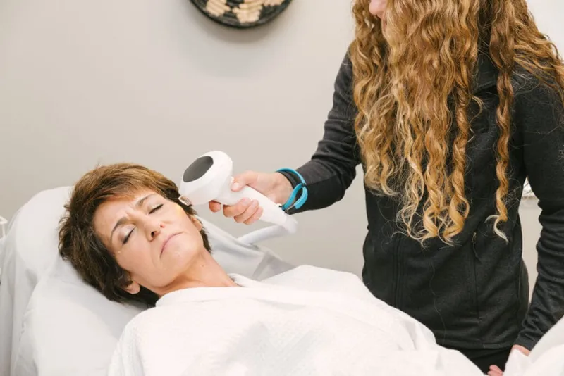 Esthetician applying a handheld device to a reclining woman's cheek during a facial treatment