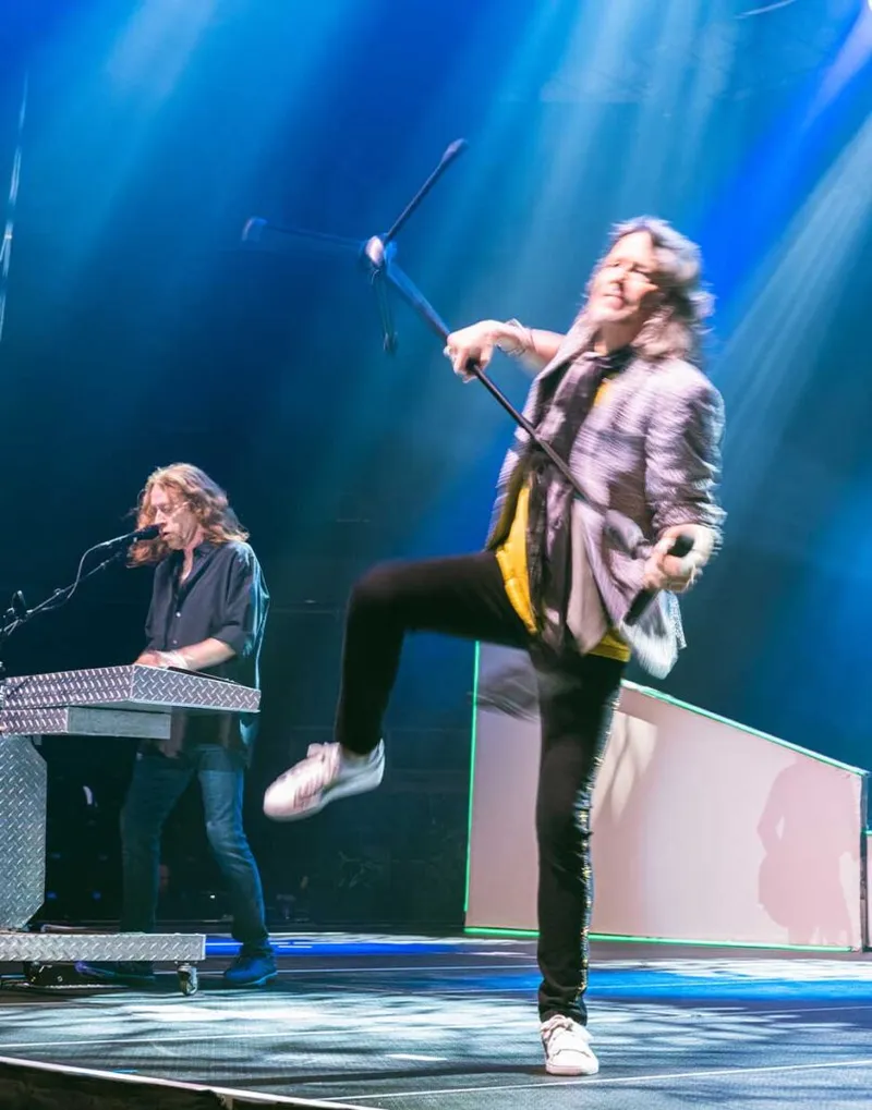 Energetic vocalist kicks leg while swinging mic stand, keyboardist performs behind him under blue stage lights