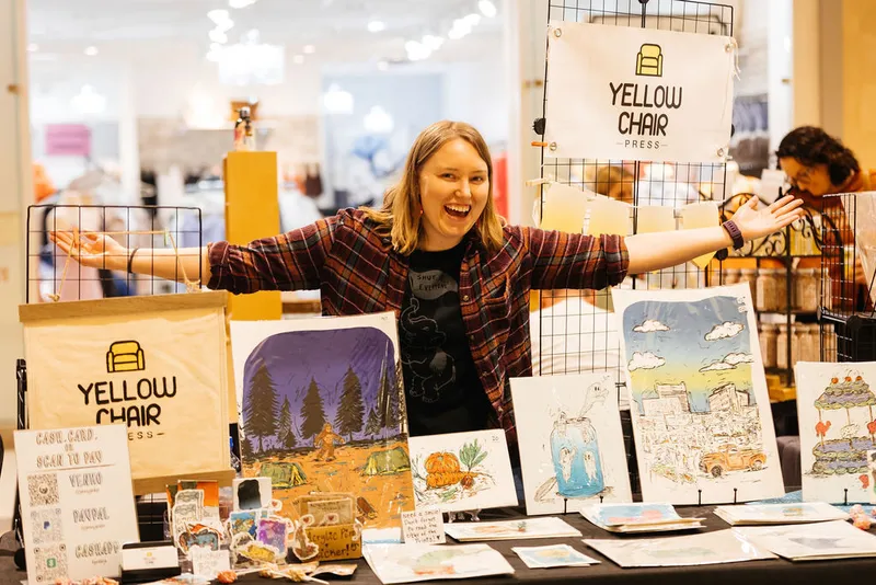 Smiling vendor spreads arms wide at a Yellow Chair Press craft fair booth displaying colorful illustrated prints