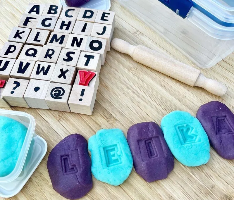 Purple and teal play dough patties stamped with letters, beside wooden alphabet stamps and a small rolling pin
