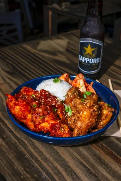 Blue bowl of saucy Korean fried wings with white rice and a Sapporo beer bottle on a wood table