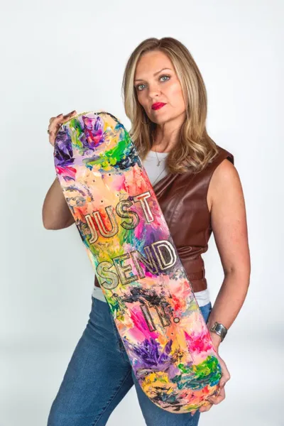 Woman in brown vest holding a colorful painted skateboard reading 'Just Send It