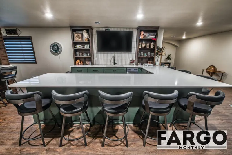 Large white quartz bar island with green cabinetry, black barstools, and a wall-mounted TV in a finished basement