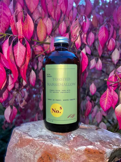 Dark glass bottle of Toasted Marshmallow simple syrup No. 8 on a rock with vivid pink autumn leaves behind it